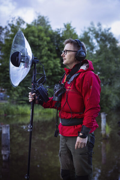 En man i röda kläder, med hörlurar håller upp en mikrofon med parabol för att fånga nattsångarnas läten.