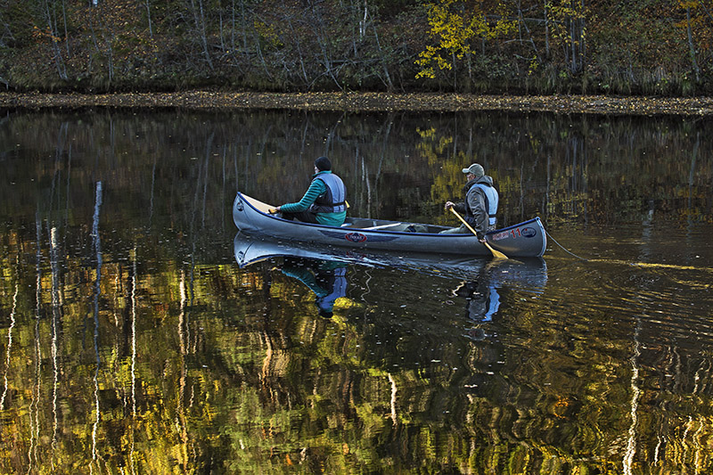 Två män paddlar i en kanot på ett stilla vatten där träd i höstfärger speglar sig.