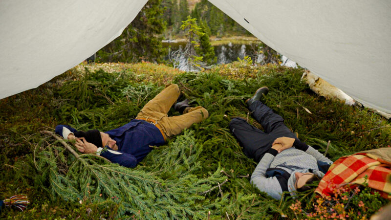 Två män ligger på granris under en tältduk under inspelningen av ett naturprogram.